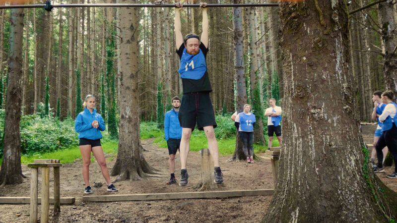 man hanging onto a poll in a tree with people watching